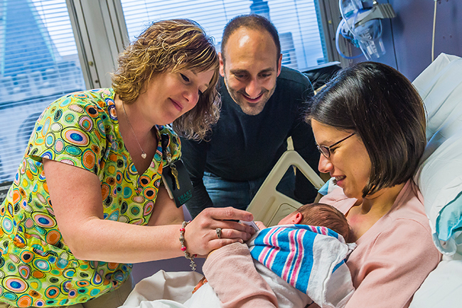A mother in hospital bed holds her newborn baby while a nurse and father look on