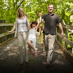 A family hiking across a wooden bridge in the woods, the father and mother lifting their smiling daughter into the air