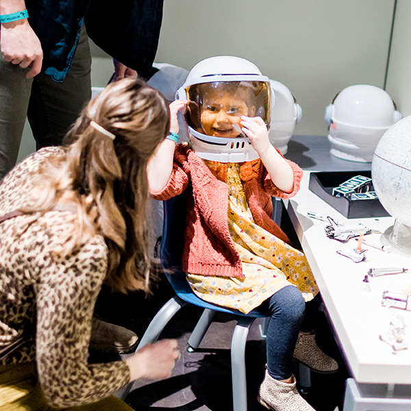 A young girl sits at a desk smiling and playing with a toy space helmet