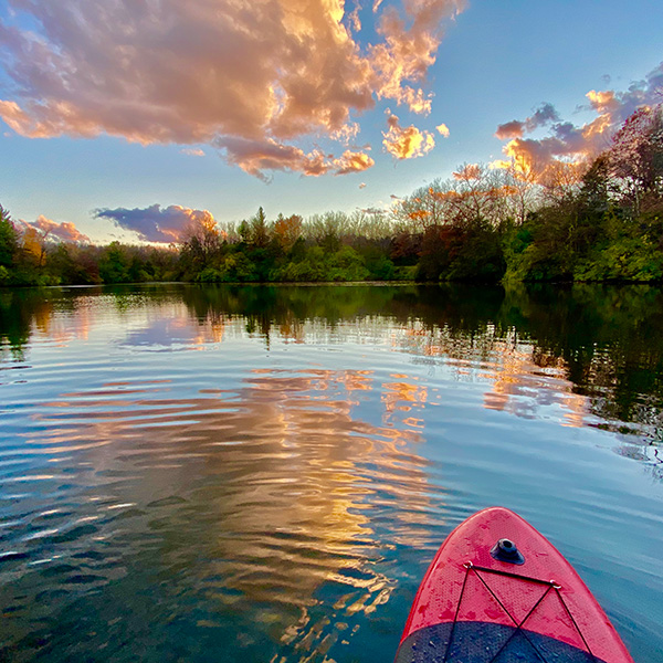 Looking on the water from the edge of a kayak with a beautiful sunset and foliage in the background.