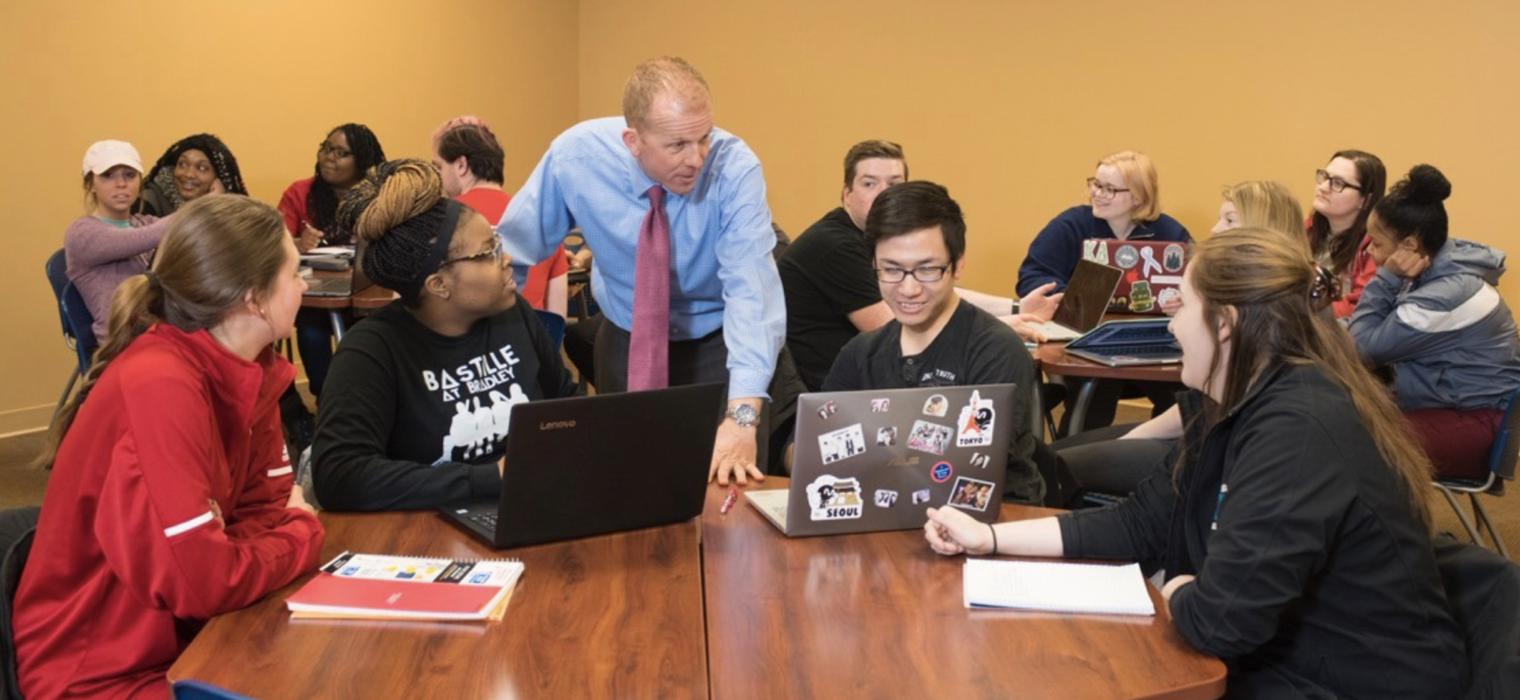 A faculty member speaks with a group of students gathered around a table with notebooks and laptops