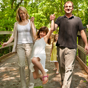 A family hiking across a wooden bridge in the woods, the father and mother lifting their smiling daughter into the air