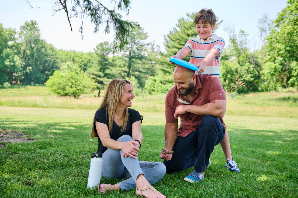 Leanne Skuse and her family laugh together in a park. Her son playfully puts a frisbee on his dad's head.